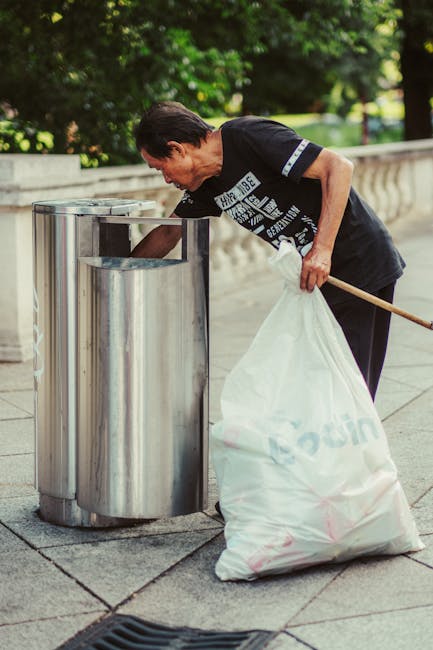 A middle-aged man with dark hair, dressed in a black T-shirt with white text and dark trousers, is seen leaning over a modern stainless steel rubbish bin on a paved sidewalk. He is holding a large white garbage bag in his left hand, which appears to be filled with waste, while his right hand is reaching into the open top of the bin. The bin features a sleek, cylindrical design with a smooth, polished finish, situated against a backdrop of lush green trees and a low stone or concrete parapet wall, suggesting an outdoor urban environment. The scene is illuminated by natural daylight, casting gentle shadows on the pavement. The overall setting emphasizes a focus on waste disposal or rubbish collection in a public space, aligning with services such as independent rubbish removal providers. The presence of the waste bag and the act of disposing waste directly support themes related to rubbish collection and waste management.