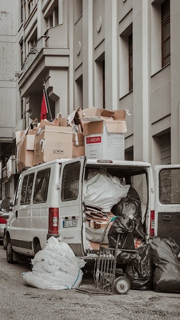 A white commercial van parked on a city street adjacent to a tall, multi-storey building with a light grey facade and rectangular windows. The van’s rear doors are open, revealing a dense load of various discarded materials including large black and white garbage bags, flattened cardboard boxes, wooden pallets, and other waste items piled inside and on the ground nearby. Several cardboard boxes, some partially collapsed or torn, are stacked on top of the vehicle, with a few marked with labels such as 'LEARDIN.' A small shopping cart filled with additional waste bags leans against the open side of the van. The scene is lit with natural daylight, highlighting the textures of the materials and the urban environment. The surroundings suggest an area where independent rubbish disposal or on-site clearance by rubbish collection services like Rubbish Collection Islington takes place, emphasizing the context of private waste handling in an urban setting.