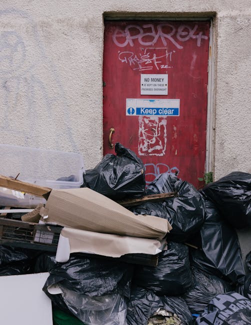 A middle-aged man with dark hair, dressed in a black T-shirt with white text and dark trousers, is seen leaning over a modern stainless steel rubbish bin on a paved sidewalk. He is holding a large white garbage bag in his left hand, which appears to be filled with waste, while his right hand is reaching into the open top of the bin. The bin features a sleek, cylindrical design with a smooth, polished finish, situated against a backdrop of lush green trees and a low stone or concrete parapet wall, suggesting an outdoor urban environment. The scene is illuminated by natural daylight, casting gentle shadows on the pavement. The overall setting emphasizes a focus on waste disposal or rubbish collection in a public space, aligning with services such as independent rubbish removal providers. The presence of the waste bag and the act of disposing waste directly support themes related to rubbish collection and waste management.