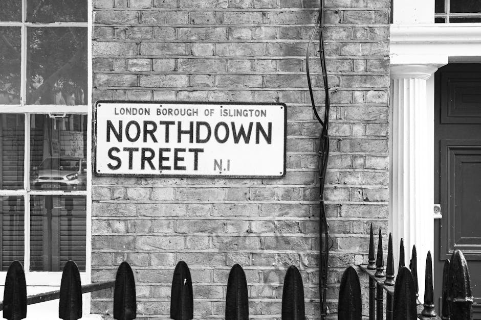 A black and white photograph of a brick building wall with a rectangular street sign affixed to it, which reads 'Northdown Street' and indicates it is within the London Borough of Islington. To the left of the sign, several small panes of a window with standard white framing are visible, with reflections of parked cars in the glass. To the right of the sign, there are visible black electrical wires running vertically along the brick wall and real black metal fence posts with pointed tops in the foreground, forming part of a small boundary fence. The brickwork shows a weathered texture with a mix of smooth and rough mortar joints, and the wall appears slightly aged. The environment suggests an urban residential setting with a typical street scene, offering an accurate depiction relevant to local rubbish removal and private waste management services available through companies like Rubbish Collection Islington, which specialize in alternative waste handling solutions beyond council curbside collections.