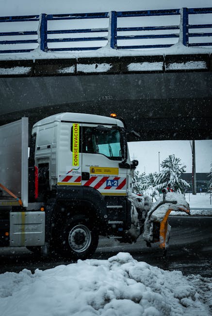 A large white snow plough vehicle equipped with a yellow and red striped plow blade is clearing snow from an urban road beneath a blue steel bridge with white guardrails. The vehicle has a high cab with a small yellow sign on the side reading 'CONSERVACION' and a blue and white label 'A-2' on the door. Snow surrounds the vehicle on the ground and partially covers parked cars visible in the background, along with snow-laden trees and overcast sky, indicating winter weather. The scene appears to be outside a waste management or rubbish collection service, illustrating an independent or private approach to maintaining road safety during winter conditions, similar to the scope of rubbish removal and clearance services provided by companies like Rubbish Collection Islington.