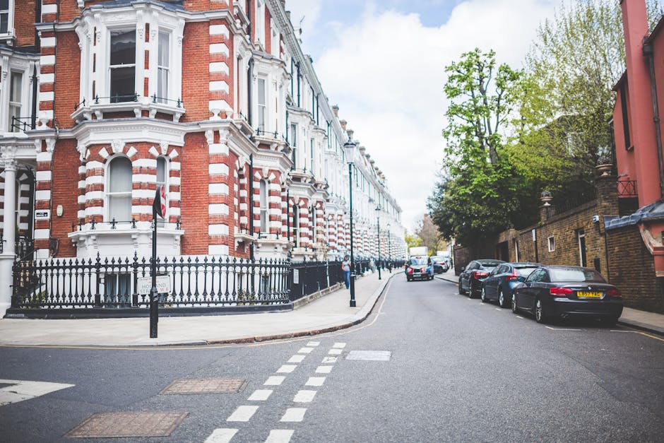 A residential street in an urban area featuring a row of historic terraced houses with Victorian architecture, constructed from red brick with white decorative trim and ornate window frames. The buildings are situated on the left side of the image, with a black wrought iron fence separating the pavement from the properties. The sidewalk runs alongside the houses, with a few small shrubs visible within the fenced areas. On the right side of the street, several parked cars, including dark-colored sedans and hatchbacks, are lined up along the curb, with some facing away from the camera. The street surface is paved with asphalt, and there are white dashed lane markings indicating directing traffic flow. Tall trees with fresh green leaves grow along the right side, providing partial shade and a natural element to the scene. In the background, a white delivery truck is partially visible down the street, while a pedestrian can be seen walking along the pavement. Overhead, the sky is partly cloudy with patches of blue, and the overall scene suggests a typical day in a busy, well-maintained neighbourhood. This setting aligns with the kind of environment where services like those offered by Rubbish Collection Islington for private waste clearance could be applicable. The image depicts an area that might require regular rubbish removal or on-site clearance services for residential or commercial waste, fitting into the context of alternative waste handling in urban environments.
