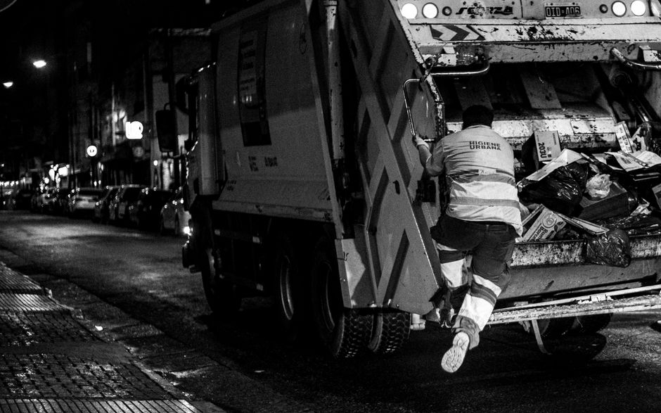 A worker wearing a high-visibility jacket with reflective stripes, marked with the words 'HIGIENE URBANA,' is in the process of unloading rubbish from a large urban waste collection truck parked on a city street at night. The worker is climbing into the truck's rear compartment, which is filled with assorted waste materials including black and clear plastic bags, cardboard boxes, and other refuse items. The truck features a hydraulic lifting mechanism, partly visible on the right side, used for compacting and emptying waste. The street scene is illuminated by streetlights, with parked cars visible along the curb in the background and storefronts faintly lit, indicating a commercial or residential area. The environment appears damp, with wet pavement reflecting the lights, emphasizing the nighttime setting. This scene illustrates the process of private waste collection and on-site rubbish removal, aligning with non-local-authority waste disposal services often employed for efficient rubbish management in urban areas, as provided by companies like Rubbish Collection Islington.