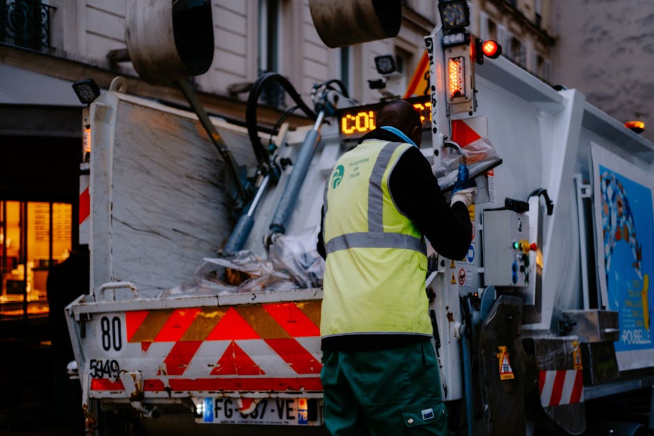 A worker wearing a high-visibility jacket with reflective stripes, marked with the words 'HIGIENE URBANA,' is in the process of unloading rubbish from a large urban waste collection truck parked on a city street at night. The worker is climbing into the truck's rear compartment, which is filled with assorted waste materials including black and clear plastic bags, cardboard boxes, and other refuse items. The truck features a hydraulic lifting mechanism, partly visible on the right side, used for compacting and emptying waste. The street scene is illuminated by streetlights, with parked cars visible along the curb in the background and storefronts faintly lit, indicating a commercial or residential area. The environment appears damp, with wet pavement reflecting the lights, emphasizing the nighttime setting. This scene illustrates the process of private waste collection and on-site rubbish removal, aligning with non-local-authority waste disposal services often employed for efficient rubbish management in urban areas, as provided by companies like Rubbish Collection Islington.