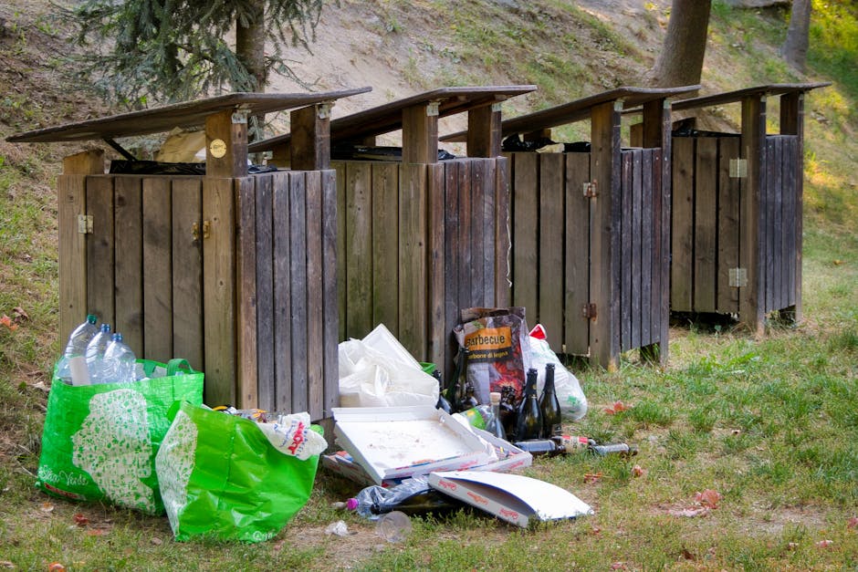 An outdoor area featuring four wooden rubbish bins with sloped roofs, positioned on a grassy patch with some scattered leaves. The bins are arranged in a row, each constructed from vertically aligned dark brown wooden planks with metal hinges and locks. In front of the bins, there are several bags and loose trash, including a large green plastic shopping bag filled with empty plastic bottles, a white foam food container, a cardboard box containing what appears to be burnt food or ashes, and a few glass bottles. The background includes a hillside with exposed soil and a few trees, suggesting a semi-natural environment. The scene appears to be a place where waste has been temporarily accumulated, with indications of casual or illegal rubbish disposal, aligning with a context where independent waste collection or on-site clearance might be necessary. Rubbish Collection Islington offers professional disposal services that could address such waste management challenges efficiently and responsibly.