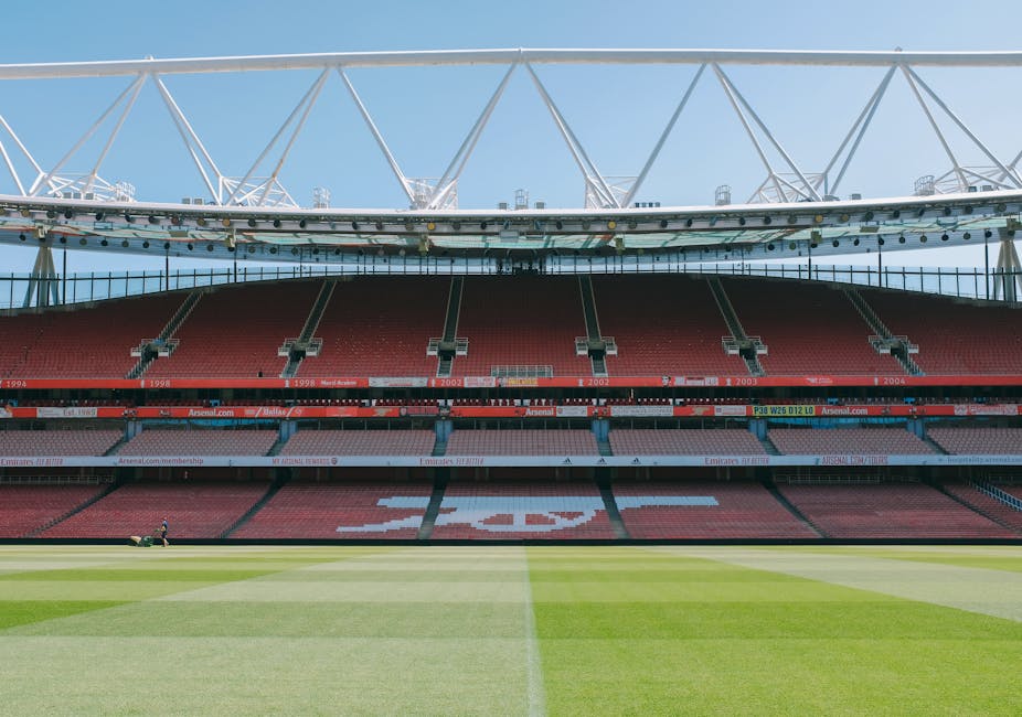 A view of the interior of a large football stadium with multiple tiers of red seating arranged in curved rows, and a partially visible pitch with neatly mowed grass in the foreground. The seating area features an Arsenal logo form created with white and red chairs. Above, a white steel framework supports a modern roof structure that extends outward, with the sky visible behind. The stadium appears empty, with no spectators present, and some small equipment or structures are seen in the background along the upper tiers. The environment is brightly lit by natural daylight, highlighting the clean, well-maintained appearance of the stadium’s seating and field. This scene exemplifies a professional sports venue, often associated with large-scale events and gatherings, and is not related to waste management or rubbish removal services.