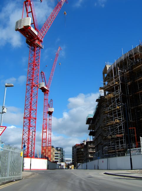 The image depicts a construction site with two large red tower cranes equipped with white counterweights and long booms extending into a blue sky with scattered clouds. To the right, there is a multi-storey building under ongoing construction, surrounded by metal scaffolding, wooden formwork, and safety barriers. The scene is situated on a paved roadway with a metal fence section visible at the foreground, indicating an active construction zone. A streetlamp is positioned on the left side of the image, and a traffic sign with partial visibility is also present. The environment appears well-lit with daylight, highlighting the industrial setting associated with on-site clearance and private waste removal activities often related to construction project waste management. The scene emphasizes the scale of building development and the importance of proper rubbish and debris management during construction phases, aligning with services like those provided by Rubbish Collection Islington for alternative waste handling solutions.