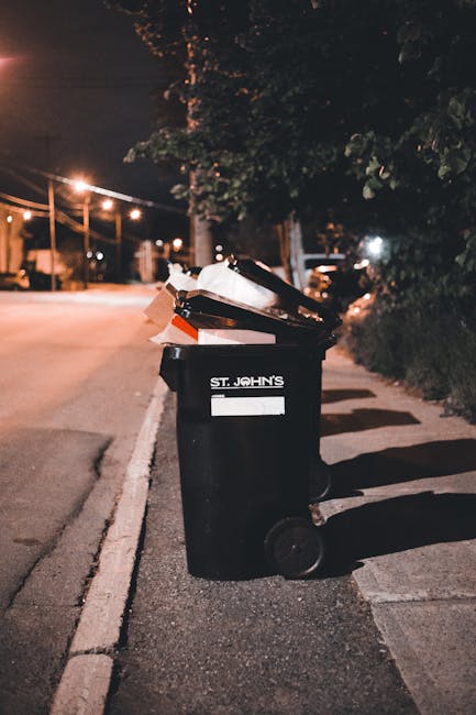 A black wheeled rubbish bin with the label 'ST. JOHN'S' is placed on the pavement beside a curb in a residential area during nighttime. The bin is filled with various waste materials, including cardboard boxes, paper, and plastic containers, some of which are partially protruding from the open lid. The bin is positioned near a row of dense bushes and trees on the right side, casting shadows on the ground. In the background, there are streetlights illuminating the quiet street, with a few parked vehicles and faintly visible houses, suggesting a suburban environment. The scene appears to depict an instance of private or independent waste collection, aligning with services offered by waste management specialists like Rubbish Collection Islington, especially for on-site rubbish clearance or alternative disposal methods outside regular municipal collections. The overall atmosphere conveys an everyday refuse disposal scenario in an urban neighborhood.