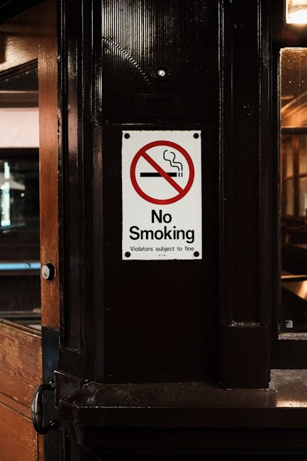 A close-up of a rectangular 'No Smoking' sign affixed to a glossy black-painted wooden or metal panel in an interior setting, possibly within a commercial or public space. The sign features a red circle with a diagonal line crossing out a black cigarette with white smoke rising from it. Below the symbol, the text reads 'No Smoking,' with a smaller line underneath stating 'Violators subject to fine.' The panel appears to be part of a partition or wall, with nearby framing visible on the left side, and reflections or faint light sources indicating a warm lighting environment. The surface has a smooth, polished finish, contrasting with the white background of the sign, ensuring clear visibility. This setting could relate to staff or public health and safety measures, aligning with the context of regulated waste or environment management within the premises, as managed by services like Rubbish Collection Islington.