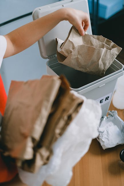 A person is seen disposing of paper waste into a white rubbish bin with a partially open lid, positioned on a wooden surface. The person's arm, wearing a light-colored sleeve, is holding a crumpled brown paper bag, poised to drop it into the bin, which is marked with a black letter on its side. In the foreground, additional crumpled brown and white paper bags are being held or are nearby, and the environment appears to be an indoor setting with soft, natural lighting. The scene reflects a typical domestic or small-scale waste disposal process, illustrating the act of segregating paper rubbish, which Rubbish Collection Islington or similar services might handle as part of their waste management operations, offering residents an alternative to on-site clearance or private disposal options. The textures include crumpled paper, smooth plastic, and the wooden surface, while the colors are subdued shades of brown, white, and neutral tones, emphasizing the practical aspect of rubbish collection and waste sorting in a residential context. All visible objects are accurately described with no assumptions about their specific use or context beyond what is shown visually.