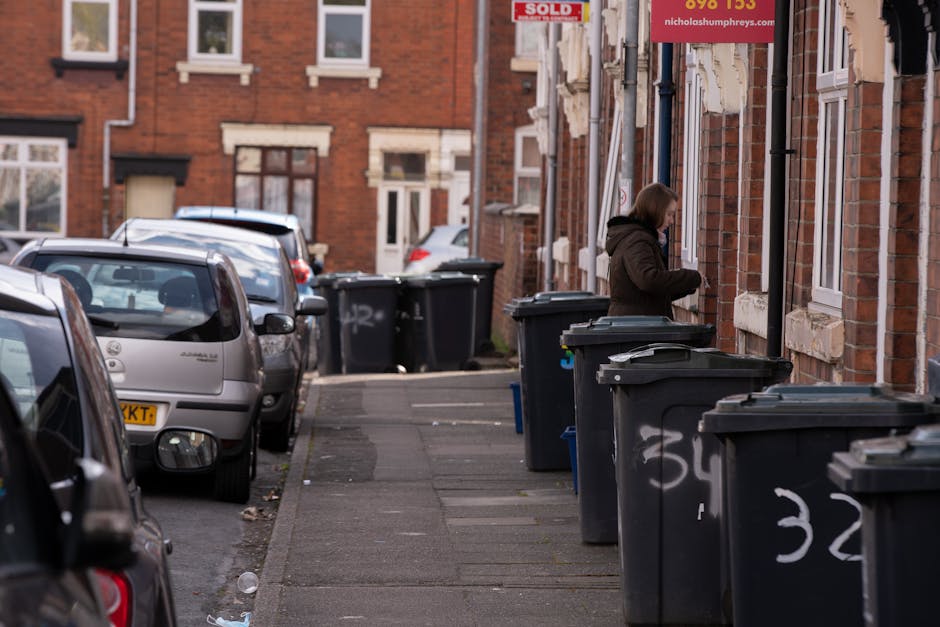A person is seen disposing of paper waste into a white rubbish bin with a partially open lid, positioned on a wooden surface. The person's arm, wearing a light-colored sleeve, is holding a crumpled brown paper bag, poised to drop it into the bin, which is marked with a black letter on its side. In the foreground, additional crumpled brown and white paper bags are being held or are nearby, and the environment appears to be an indoor setting with soft, natural lighting. The scene reflects a typical domestic or small-scale waste disposal process, illustrating the act of segregating paper rubbish, which Rubbish Collection Islington or similar services might handle as part of their waste management operations, offering residents an alternative to on-site clearance or private disposal options. The textures include crumpled paper, smooth plastic, and the wooden surface, while the colors are subdued shades of brown, white, and neutral tones, emphasizing the practical aspect of rubbish collection and waste sorting in a residential context. All visible objects are accurately described with no assumptions about their specific use or context beyond what is shown visually.