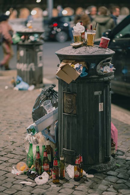 A black metal public waste bin with a domed lid is situated on a cobbled street, with various discarded items surrounding it. The lid is slightly open, and on top of it, there are several takeaway beverage cups, a plastic cup with a straw, and a small cardboard container, all containing or associated with food or drink waste. Attached to the side of the bin is a crumpled cardboard box, and a black plastic rubbish bag is tied nearby. The ground around the bin is littered with glass bottles, mainly green and brown, some of which are tipped over or partially crushed, along with paper wrappers, tissues, and other small pieces of waste. In the background, blurred figures of pedestrians and parked cars are visible, with a grey plastic bin further back, all under soft, natural daylight, suggesting an urban environment where private or independent rubbish disposal might be required. Rubbish Collection Islington services such waste management in the area, highlighting the importance of proper waste disposal and cleaning of public spaces.