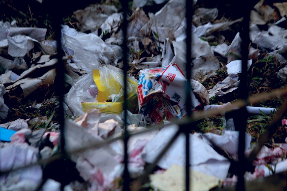 A close-up view of discarded rubbish behind a black metal fence, featuring various crumpled plastic bags, some with visible branding or printed text, among scattered paper waste and torn paper packaging. The waste appears to be mixed, with materials such as thin plastic and paper, some of which are stained or weathered. The debris is piled on the ground, partially obscured by the fence bars, in an outdoor setting, possibly a neglected area or a site awaiting rubbish removal. The environment has subdued lighting, indicating an overcast day or shaded location, highlighting the unkempt state of the litter. This scene exemplifies the type of waste that professional rubbish collection services, like those offered by Rubbish Collection Islington, might handle as part of their on-site clearance and private waste disposal solutions, supporting alternative rubbish removal methods outside standard council collection processes.