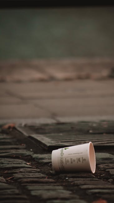 A black metal public waste bin with a domed lid is situated on a cobbled street, with various discarded items surrounding it. The lid is slightly open, and on top of it, there are several takeaway beverage cups, a plastic cup with a straw, and a small cardboard container, all containing or associated with food or drink waste. Attached to the side of the bin is a crumpled cardboard box, and a black plastic rubbish bag is tied nearby. The ground around the bin is littered with glass bottles, mainly green and brown, some of which are tipped over or partially crushed, along with paper wrappers, tissues, and other small pieces of waste. In the background, blurred figures of pedestrians and parked cars are visible, with a grey plastic bin further back, all under soft, natural daylight, suggesting an urban environment where private or independent rubbish disposal might be required. Rubbish Collection Islington services such waste management in the area, highlighting the importance of proper waste disposal and cleaning of public spaces.